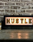 The Faire Lighted Sign featuring "HUSTLE" in bold black letters rests on a wooden surface by a brick wall, alongside stacked books and a white cup with saucer.