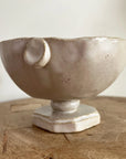 The Faire Tiny Trophy Bowl, a handmade glazed stoneware trinket dish with an off-white finish, irregular rim, small handle detail, and hexagonal pedestal base, rests on a wooden surface against a plain background.