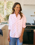 A woman with long brown hair, smiling in a bright kitchen with white tile and a green stove, wears the Frank & Eileen Eileen 26 relaxed fit shirt featuring red heart patterns and blue jeans.