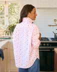 A woman stands in a sunlit kitchen, back turned, wearing the Frank & Eileen Eileen 26 relaxed shirt with tiny red hearts and blue jeans. A stove and plant complete the casual California vibe.