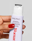A hand with red nail polish holds Faire's Let's Neck - Neck Serum w/ Cooling Applicator, topped with white lotion. The light-colored bottle features measurement markings and partial text in red and black.
