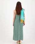 A woman with long brown hair, seen from behind, wears the Me369 Olivia Camisole Dress in green print, gold flats, and has a large turquoise and yellow scarf draped over her left shoulder.