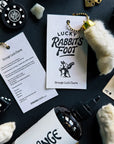 A flat lay of Faire’s Lucky Rabbit's Foot keychains tagged “Strange Luck Charm,” surrounded by dice, poker chips, cards, and a “STRANGE” bottle—the perfect good luck charm for gamblers.