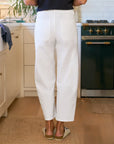 A person stands in a kitchen wearing the Frank & Eileen Everyday Barrel Pant in white and beige slip-on shoes. The kitchen has a green stove, white cabinets, and wooden floors.