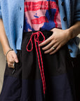 Wearing a blue cardigan, a red and blue graphic tee, and the Ping Pong Surf Club Le Vintage Sport Skirt with red rope belt, this look—completed by ring and bracelet—captures signature Ping Pong Surf Club style.