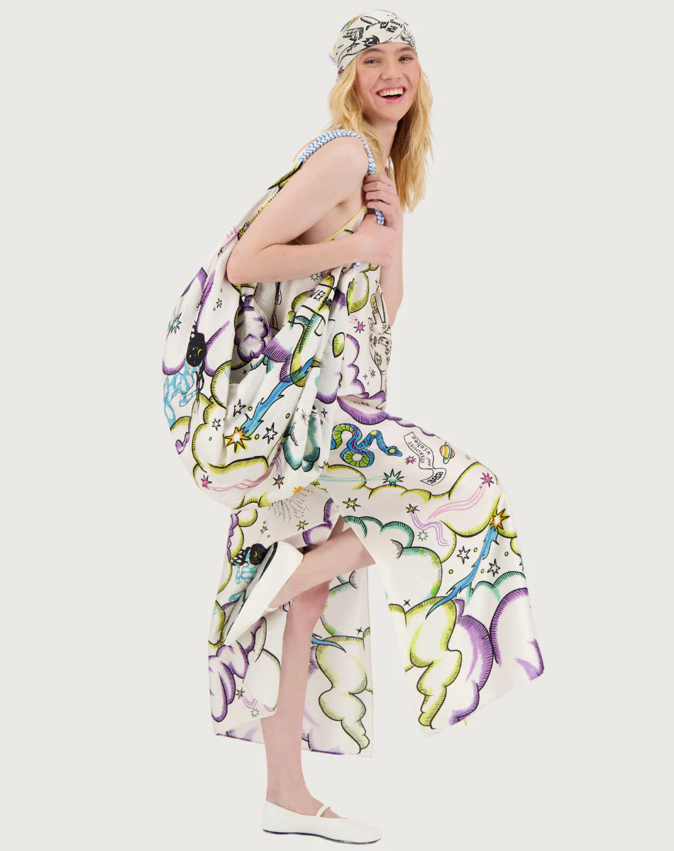 A smiling woman with long blonde hair wears the Olivia Camisole Dress by Me369 in a colorful pattern, paired with a matching headscarf and white flats. She playfully lifts one leg and looks at the camera against a plain white background.