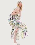 A smiling woman with long blonde hair wears the Olivia Camisole Dress by Me369 in a colorful pattern, paired with a matching headscarf and white flats. She playfully lifts one leg and looks at the camera against a plain white background.