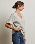 A woman with brown hair in a low bun, wearing the Perfectwhitetee French Terry SS Crew in light gray and blue jeans, stands in profile against a plain off-white background.