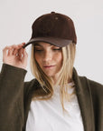 A person with long blonde hair wears the Faire Aven Leather Ball Cap, styled with an olive green jacket and white shirt, looking downward while touching the cap’s brim against a plain light background.