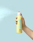A hand with light blue nails holds a yellow can of Faire Bask Mist Sunscreen, spraying a fine, reef-safe mist against a light blue background.
