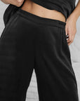 A person wears the Faire Lunya Washable Silk Pant—loose-fitting black silk with subtle side slits—styled with a black top that reveals part of their midriff, posing in front of a white bed and wall.