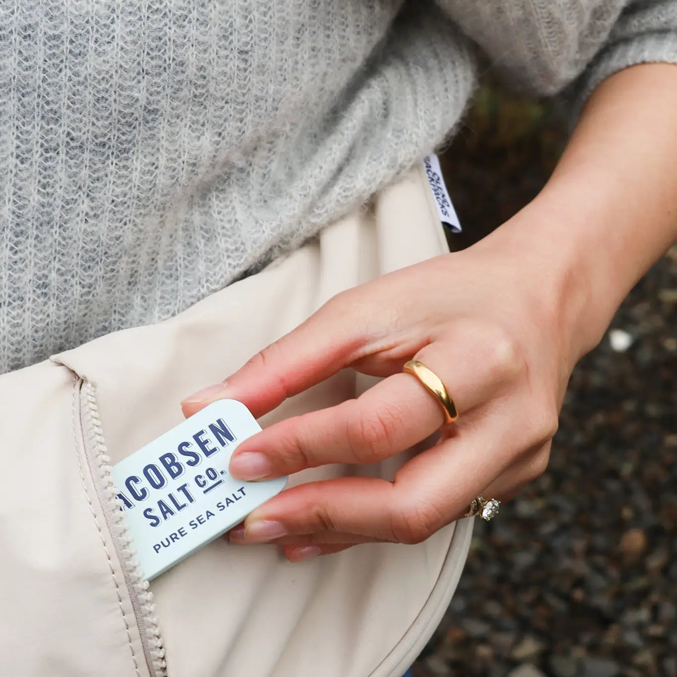 A person in a gray knit sweater pulls a "Faire Kosher Sea Salt slide Tin" from the pocket of a light-colored bag while wearing a gold ring.