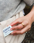 A person in a gray knit sweater pulls a "Faire Kosher Sea Salt slide Tin" from the pocket of a light-colored bag while wearing a gold ring.