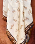 The Faire Wild Rag Silk Scarf is cream-colored with brown and tan cowboy and horse designs, a dark brown border with dashed white lines, and is styled here against a rustic metal backdrop.
