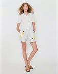 A woman with curly hair wears Kule’s The Limon—white and grey striped shorts with lemon embroidery—styled with a white cotton polo and brown sandals, standing against a plain white background.