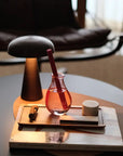 A small glass with a red drink and straw sits on a wooden tray beside the Faire Boost Reed Diffuser, atop stacked books next to a modern black lamp on a round table, with a leather chair in the background.
