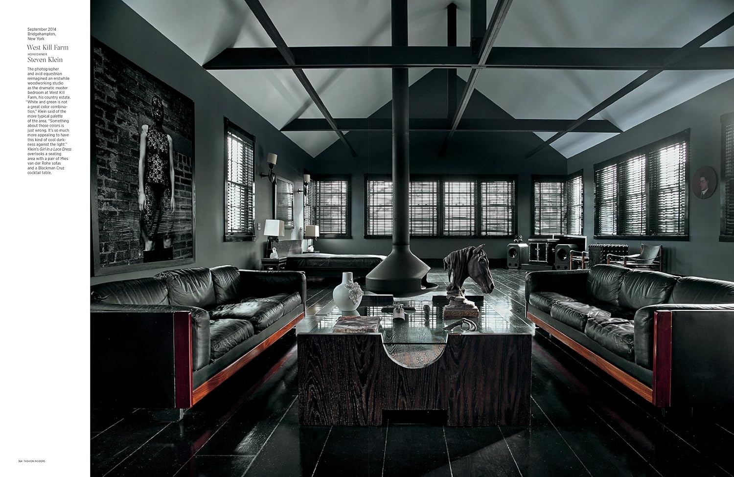 A modern living room with dark walls and floors, two black leather sofas, a central fireplace, large windows with blinds, and chic décor echoing the style of Architectural Digest at 100 by Hachette Book Group. Text appears along the left edge.