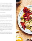 A vibrant plate of marinated peppers, cheese, olives, and artichokes sits beside Hachette Book Group’s "Make More with Less," featuring simple recipes and tips for budget-friendly cooking on a wooden table.