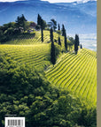 Rows of vineyards on a sunlit hillside with cypress trees and a small house, set against forested slopes and snow-capped peaks, evoke the spirit of "150 Vineyards You Need" by Simon & Schuster Inc., perfect for wine lovers.