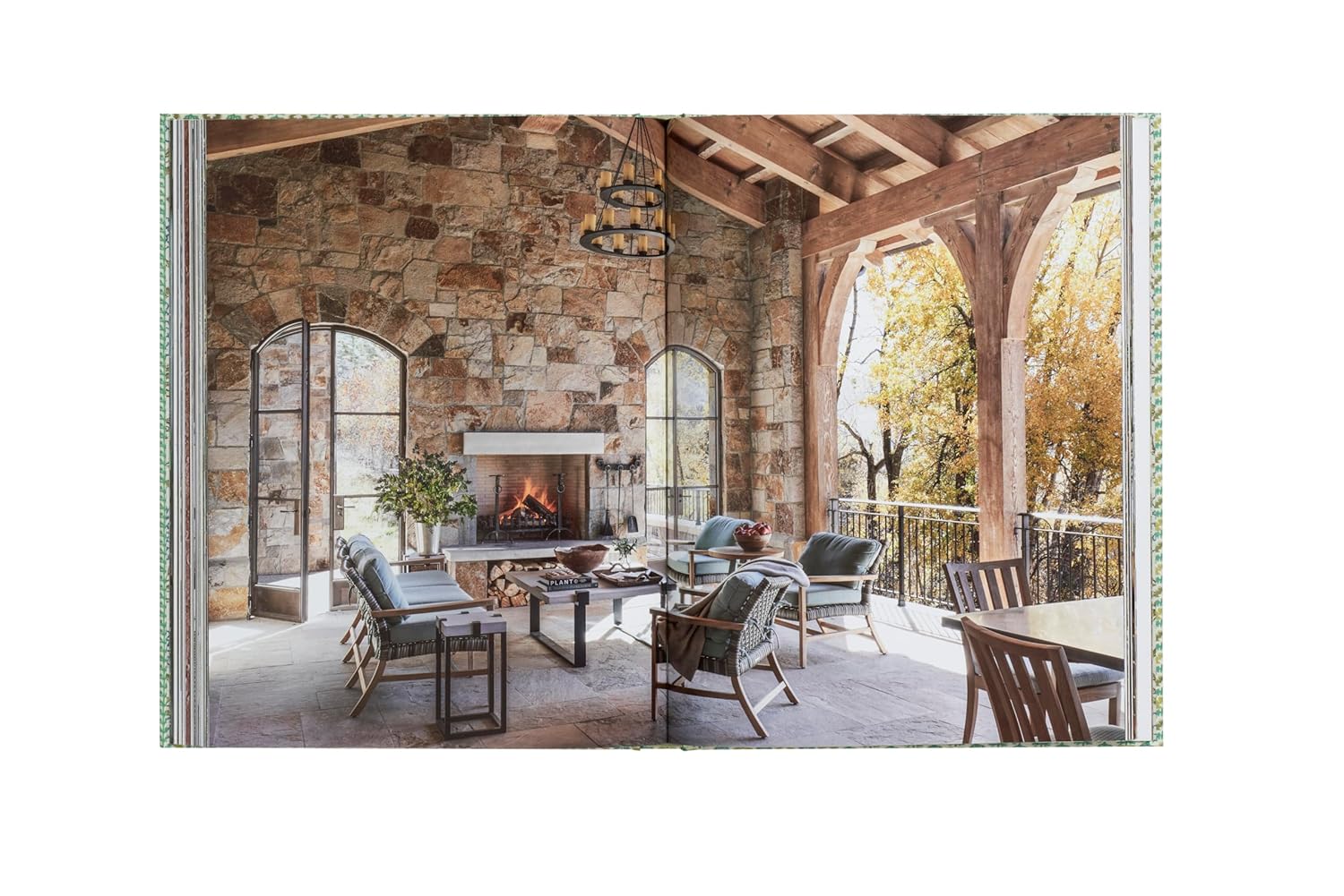 Experience well-being on a sunlit stone patio with natural materials, wooden beams, arched windows, and cozy outdoor furniture from Natural Living by Design by Hachette Book Group—designed to overlook lush trees.