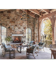 Experience well-being on a sunlit stone patio with natural materials, wooden beams, arched windows, and cozy outdoor furniture from Natural Living by Design by Hachette Book Group—designed to overlook lush trees.