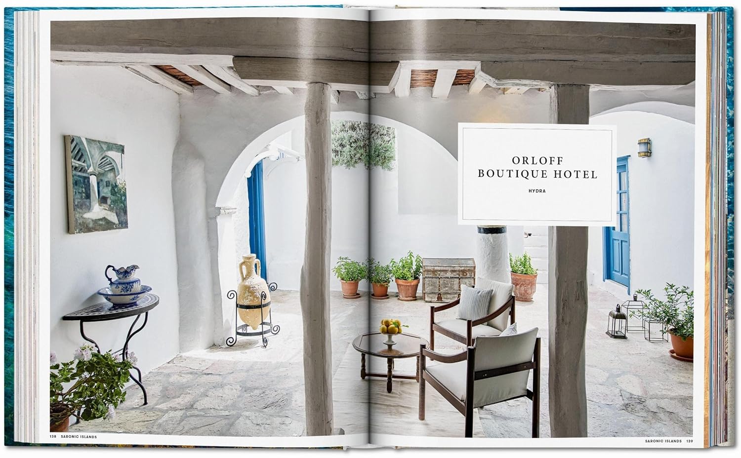 A magazine spread from "Great Escapes Greece The Hotel Book" by Ingram Book Company showcases a bright courtyard at Orloff Boutique Hotel, with potted plants, rustic beams, blue doors, and Mediterranean decor for a peaceful retreat.