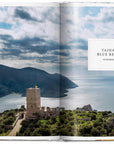 A scenic coastal view from Great Escapes Mediterranean by Ingram Book Company highlights Tainaron Blue Retreat’s stunning stone tower atop a hill overlooking the sea, mountains, and blue water beneath a partly cloudy sky.