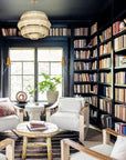 A cozy home library inspired by Hachette Book Group’s "Down to Earth," with floor-to-ceiling bookshelves on dark walls, sunlight from large windows, four white armchairs, a rustic wood table, and a woven chandelier.
