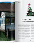 An open "New York Times Cultured Traveler 100 Trips" book by Ingram Book Company displays a lakeside pavilion photo, an illustration of a woman with mountains and lake, and an article titled "Seeing Like Klimt on a Shimmering Lake.
