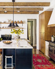 A modern kitchen featuring exposed beams, white walls, and black cabinetry channels the laid-back style of Hachette Book Group’s Down to Earth, with a large island, bar stools, pendant lights, sunflowers, and a red rug for warmth.