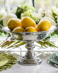 The Faire Vintage 1795 Pedestal Bowl, handmade in Italy, displays vibrant yellow lemons on a marble table accented with green plates, a matching napkin, and white flowers in the background.