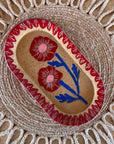 The Faire Blossom Sgraffito Dish, an oval ceramic piece with two red flowers and green stems bordered by a red pattern, is displayed on a woven sunburst placemat.