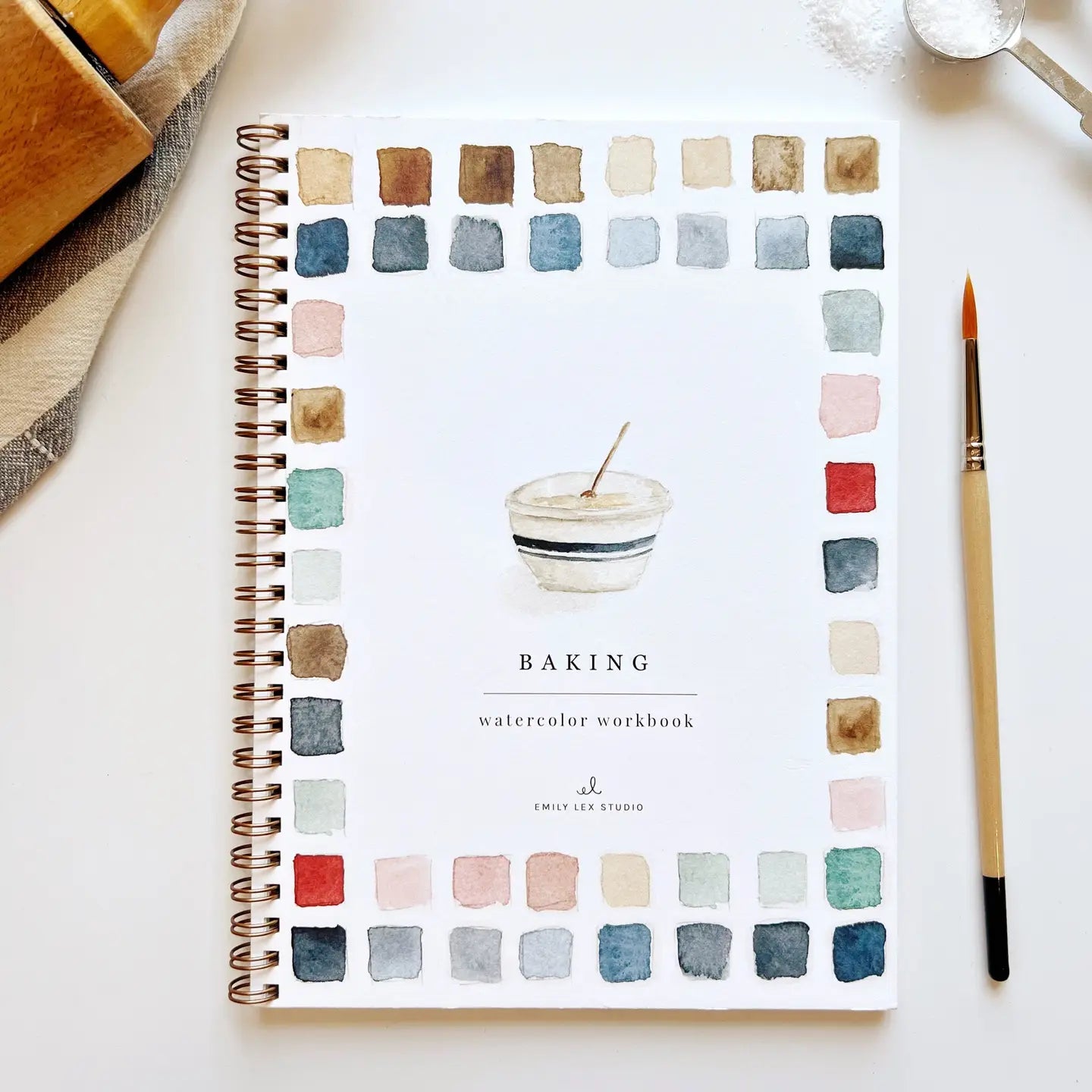 A spiral-bound "Watercolor Workbook" by Faire titled "Baking" lies on a white surface. The cover shows a bowl illustration framed by colorful paint swatches, with a paintbrush and baking items nearby, ideal for anyone wanting beginner-friendly illustrations and step-by-step instructions.