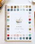 A spiral-bound "Watercolor Workbook" by Faire titled "Baking" lies on a white surface. The cover shows a bowl illustration framed by colorful paint swatches, with a paintbrush and baking items nearby, ideal for anyone wanting beginner-friendly illustrations and step-by-step instructions.