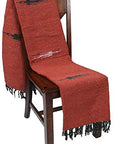 A wooden chair draped with the Faire Rust Red Baja Thunderbird Mexican Blanket. Handmade in Mexico, it features black fringe at the ends and subtle black and white patterns.