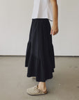 A person stands indoors in a white shirt, beige slip-on shoes, and The Ranch Skirt by Hey Gang—a long black elastic waist skirt. The side profile is shown from shoulder to feet against a plain wall.