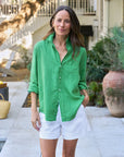 A woman stands outdoors in the Frank & Eileen Eileen 26, a bright green relaxed-fit cotton shirt with bust-flattering buttons and rolled sleeves, paired with white shorts. She smiles gently, greenery and a clay pot in the background.