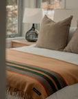 A cozy, sunlit bedroom features a neatly made bed with beige pillows, a vintage-inspired Faire S&A Blanket with green and orange stripes, and a bedside table topped by a black lamp.