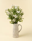 A white ceramic pitcher holds green leafy stems and Faire's 1-9528 Locust Flower Branch, creating an elegant accent against a plain light beige background.