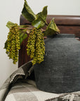 The Indaba Dripping Seed Stem, with clusters of green seed pods and dry leaves, cascades from a dark ceramic vase set on a striped neutral cloth against wood—an elegant botanical accent.