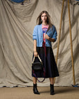 A woman stands before a beige draped backdrop, wearing a blue cardigan over a pink graphic tee, the Ping Pong Surf Club Le Vintage Sport Skirt, black ankle boots, and holding a black woven handbag with a chunky strap.