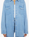 A person wears "The Exes And The Ohs" oversized denim shirt in vintage-blue wash by Mother, featuring two chest pockets and silver buttons. The Western-inspired shirt is open over a white top and paired with blue jeans, captured from shoulders to thighs.