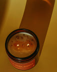 Brown jar Gold Dust Woman Candle by Faire with flower petals on a yellow surface, casting a long shadow.