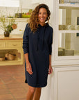 A smiling woman with curly hair wears the Frank & Eileen Mini James Hoodie Cape Dress, a relaxed fit navy blue mini dress, standing indoors by a white shelf in a bright kitchen with plants and wooden decor.
