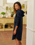 A woman with curly hair wears the Frank & Eileen Mini James Hoodie Cape Dress in navy blue, standing sideways in a cozy kitchen with cream cabinets and terracotta tiles. She looks back and gently smiles at the camera.