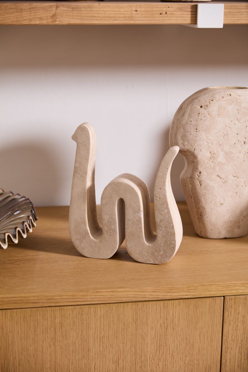 The Kingsnake Sculpture by Accent Decor sits on a wooden cabinet beside a rounded stone and metal decor, with shelves above against a white wall, adding an artistic touch to your home.