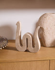 The Kingsnake Sculpture by Accent Decor sits on a wooden cabinet beside a rounded stone and metal decor, with shelves above against a white wall, adding an artistic touch to your home.