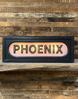 The Faire Lighted Sign, featuring "PHOENIX" in bold black letters, sits on a rustic wooden shelf against a plank wall—a striking personalized decor piece.