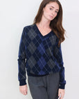 A woman with long brown hair models the Brazeau Tricot Argyle V-Neck in blue and gray, paired with gray pants, standing before a white background and facing the camera with a neutral expression.
