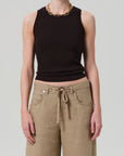 Wearing the Isabel Rib Tank by Citizens Of Humanity/AGOLDE in black, paired with tan wide-leg pants with a tie waist, a gold chain necklace, and rings—an ideal layering piece for versatile styling against a light gray background.
